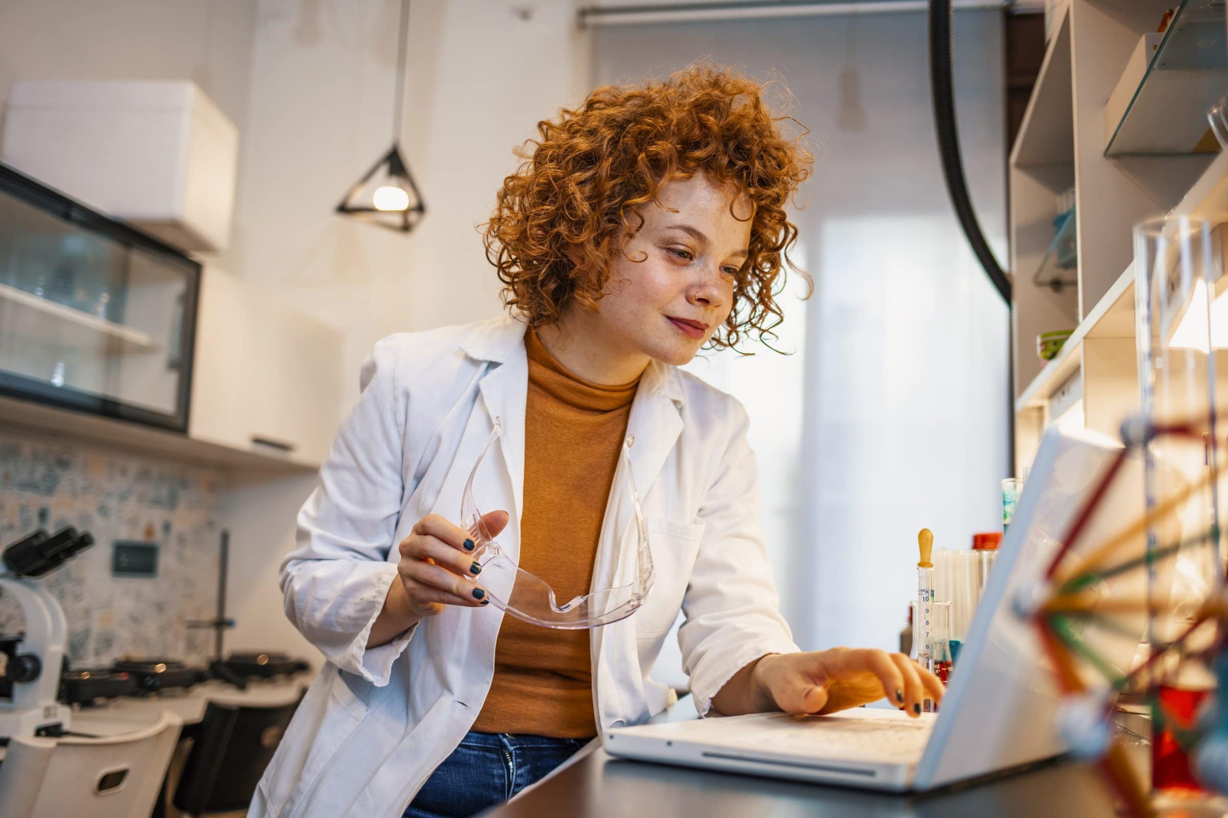 Test results. Serious smart female biologist sitting at the table and looking at the laptop screen while studying test results. Young Caucasian female scientist using laptop while sitting with microscope at desk in laboratory