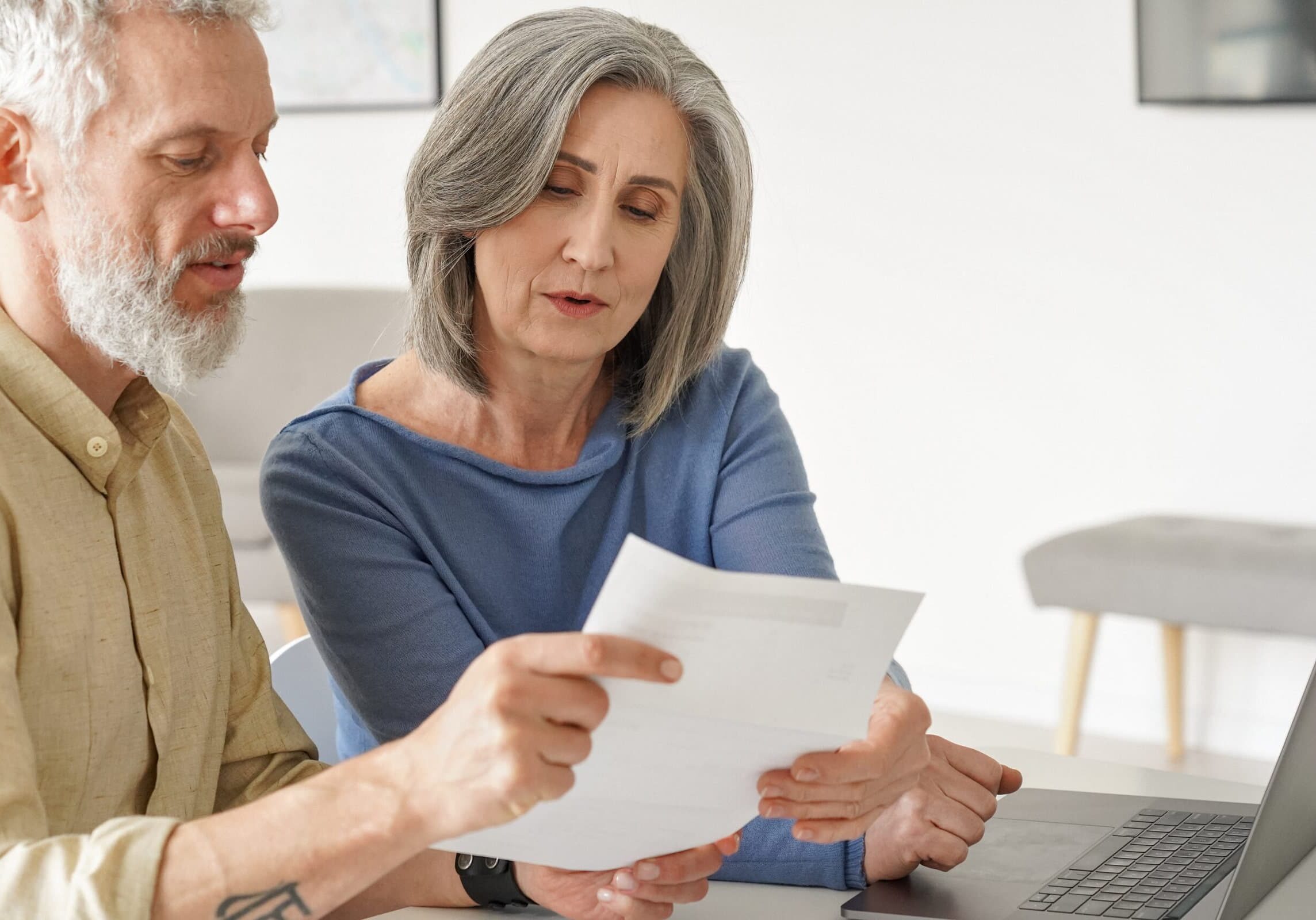 Older mature couple checking bank documents using laptop at home. Senior mid age retired man and woman reading paper bills, calculating pension or taxes, planning retirement finances, doing paperwork.