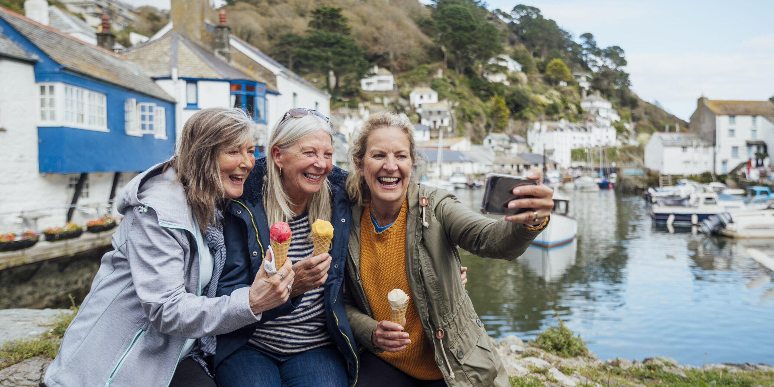 Female friends sitting on a wall and taking a selfie together on a mobile phone while eating an ice cream in Polperro, Cornwall. The harbour is behind them.