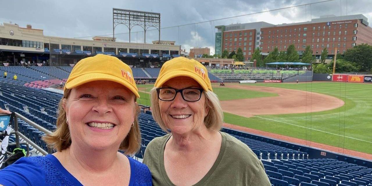 Two P.E.O.s in the stands at a baseball park wearing yellow P.E.O. ball caps