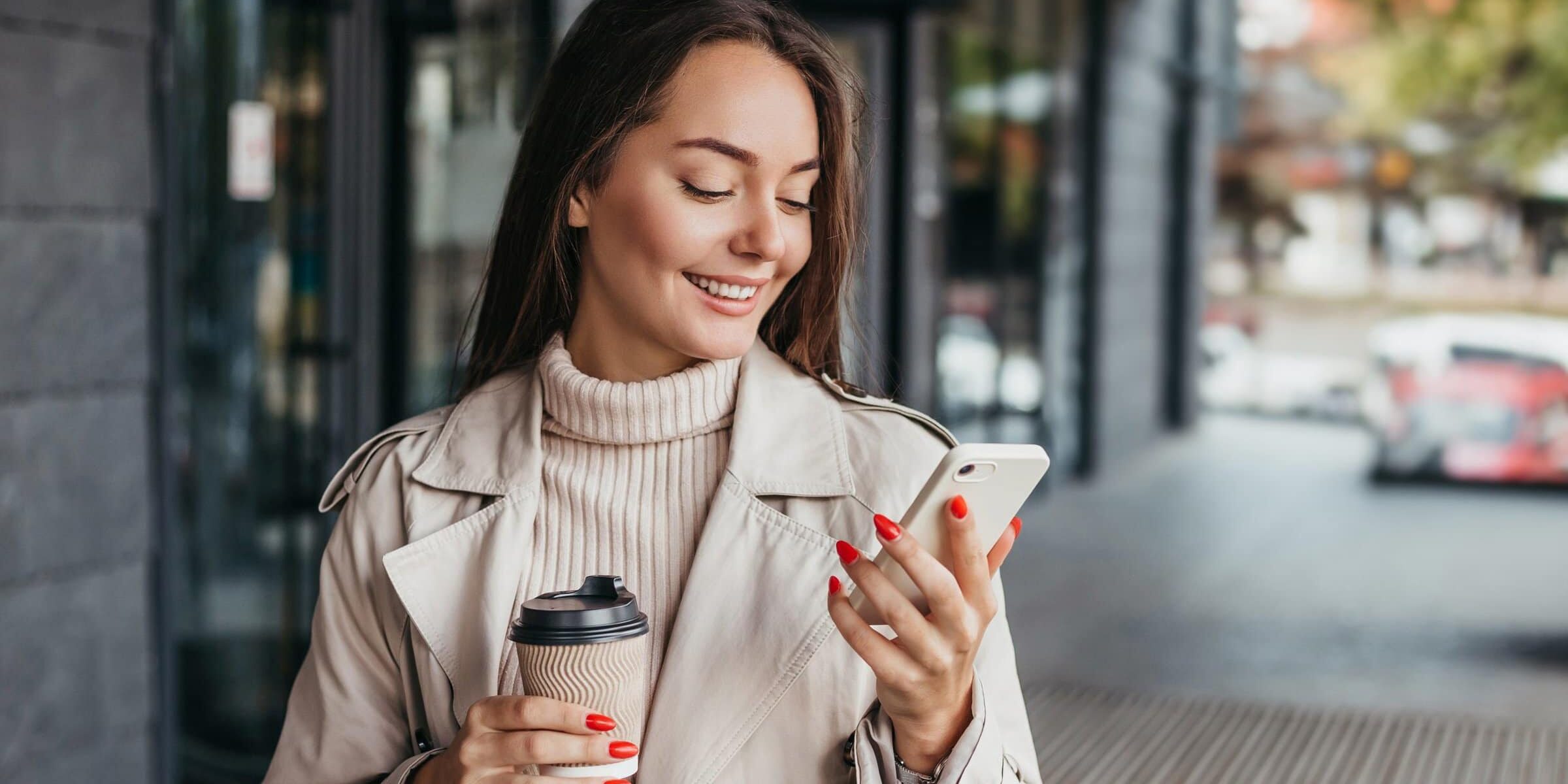Young smiling ukrainian brunette woman holding a smartphone and a cup of coffee looking at device screen against the background of an office building. copy space