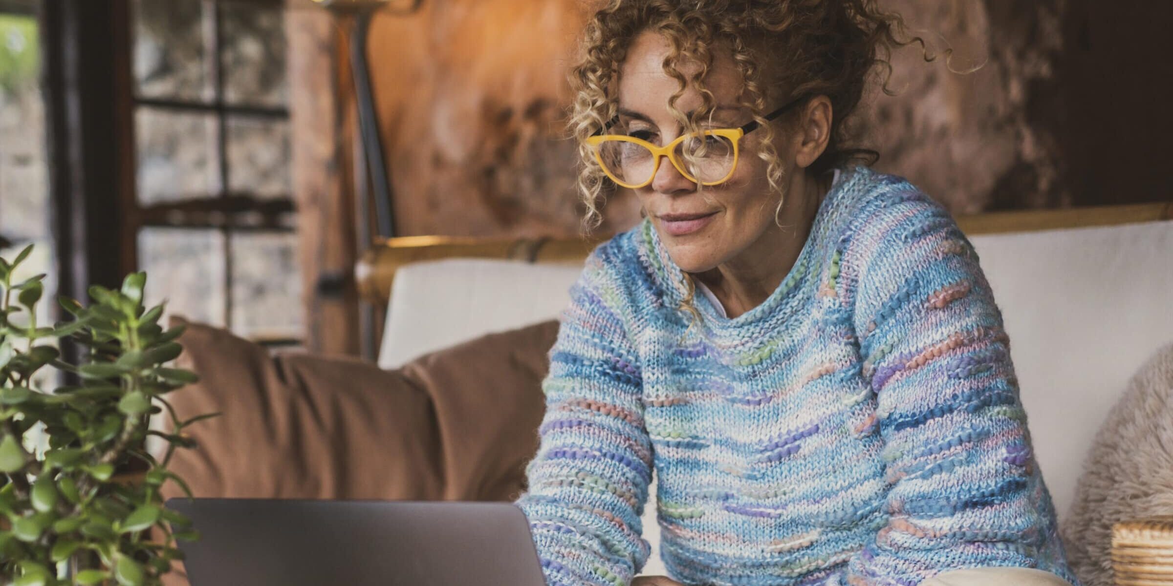 Portrait of happy modern woman at home sitting on the sofa using computer and wearing eyewear. Female people surf the web and work with laptop smiling. Indoor technology leisure activity concept